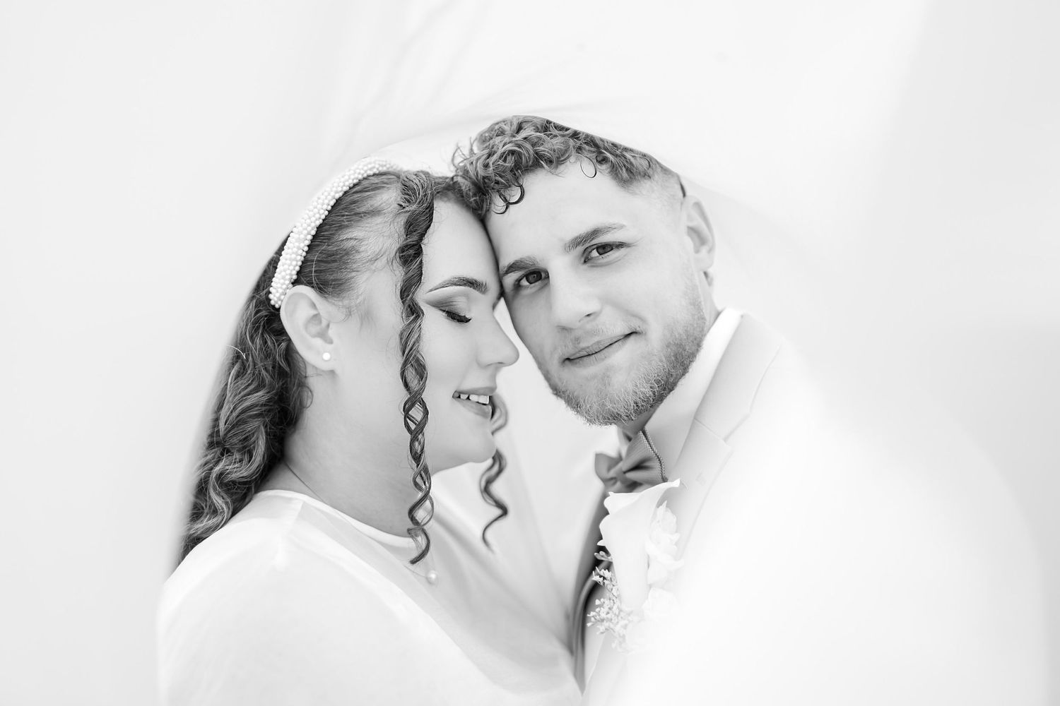 Black and white wedding portrait of a smiling bride with curly hair and a groom in a bow tie, framed by soft white veil.