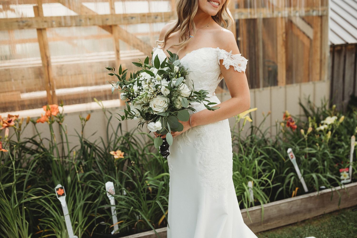 Bride in an off-the-shoulder lace wedding dress holding a white and green bouquet beside a greenhouse.