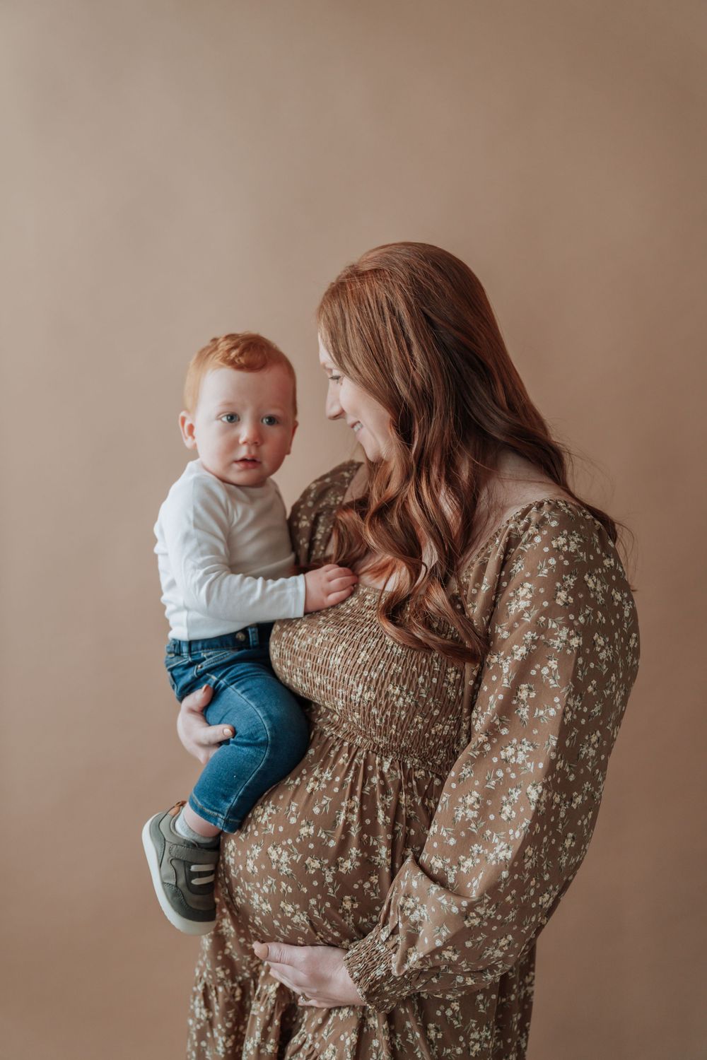 Pregnant redheaded mother in brown floral dress gazes lovingly at red-haired toddler son during studio maternity session.