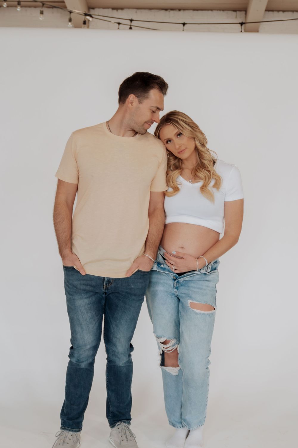Couple maternity photo, man in beige shirt and woman in white crop top with ripped jeans, smiling.