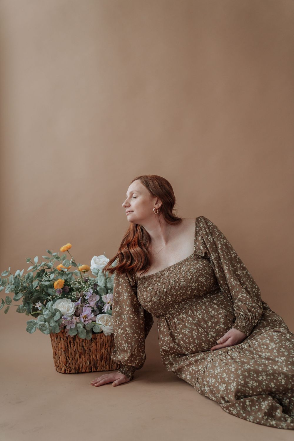 Pregnant redheaded woman in olive floral dress sits on floor resting beside wicker basket of colorful blooms.