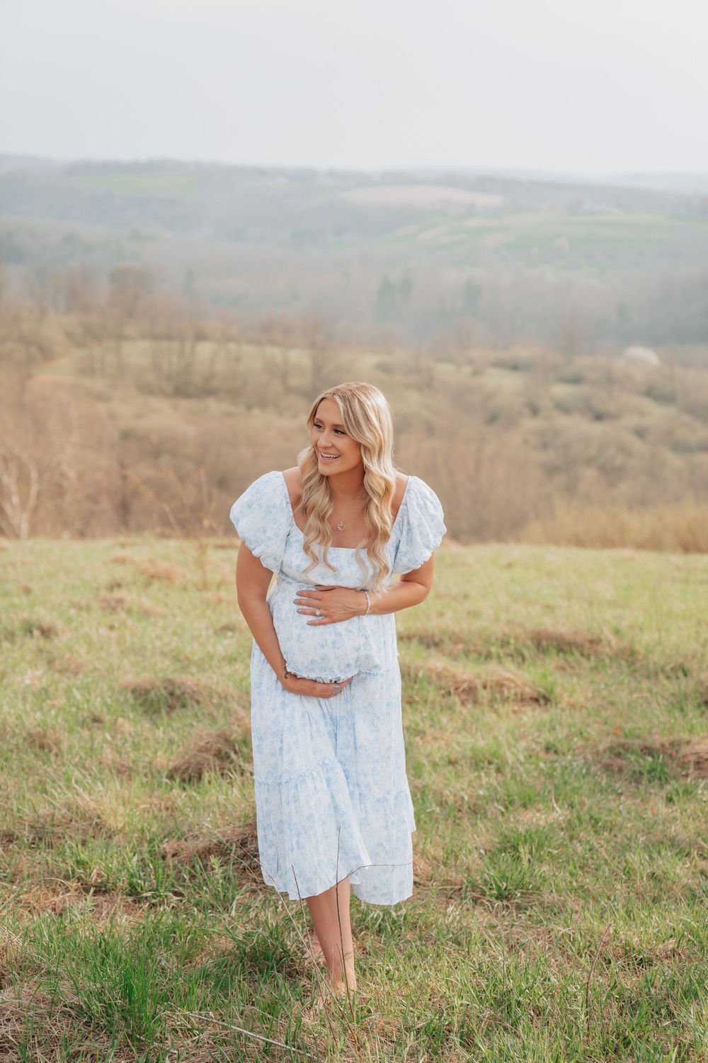 Pregnant woman in light blue floral dress holding baby bump in green field.