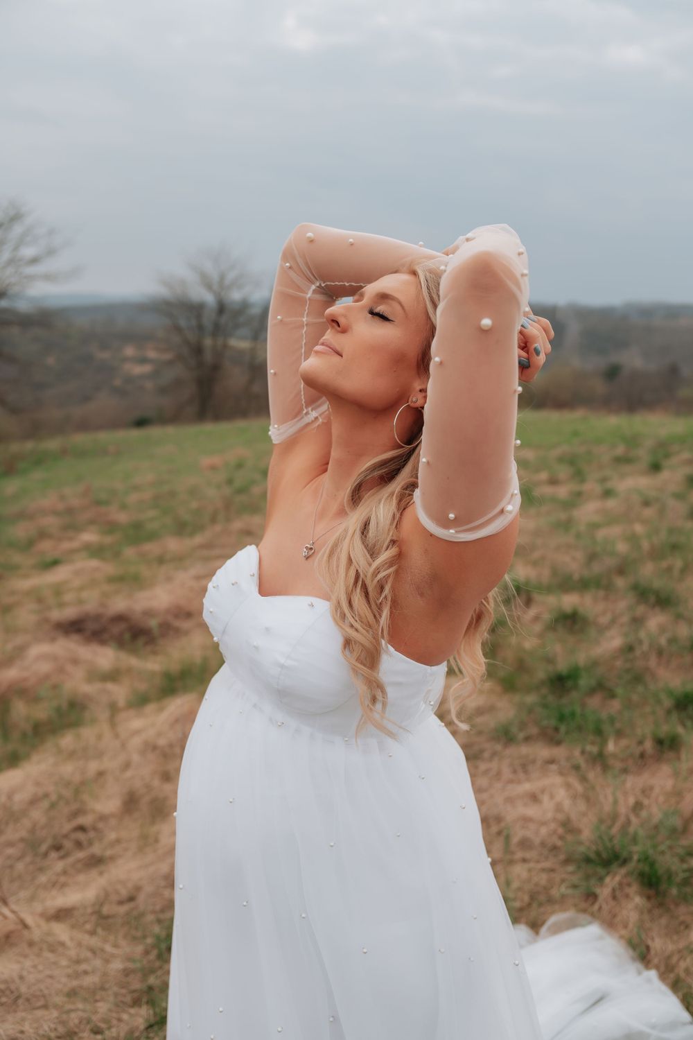 Pregnant woman in white gown with sheer pearl sleeves posing outdoors.