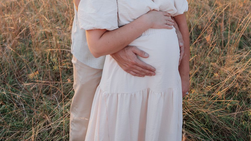 Man tenderly holding pregnant woman's baby bump from behind in warm golden field at sunset.