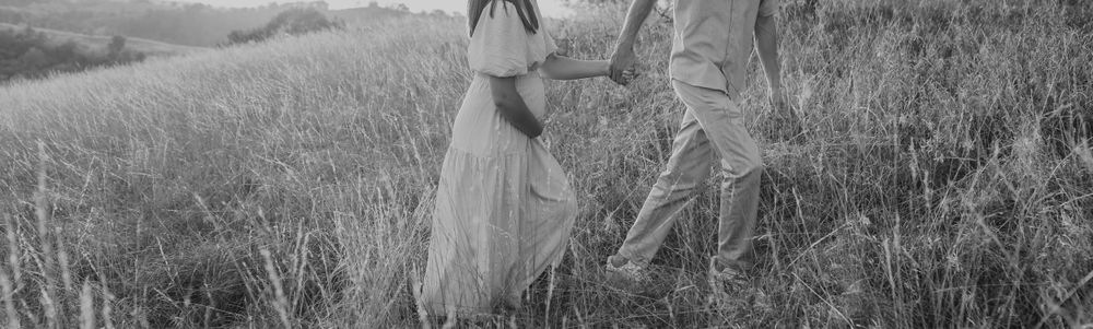 Black and white photo of a pregnant couple holding hands while walking through a lush green field.