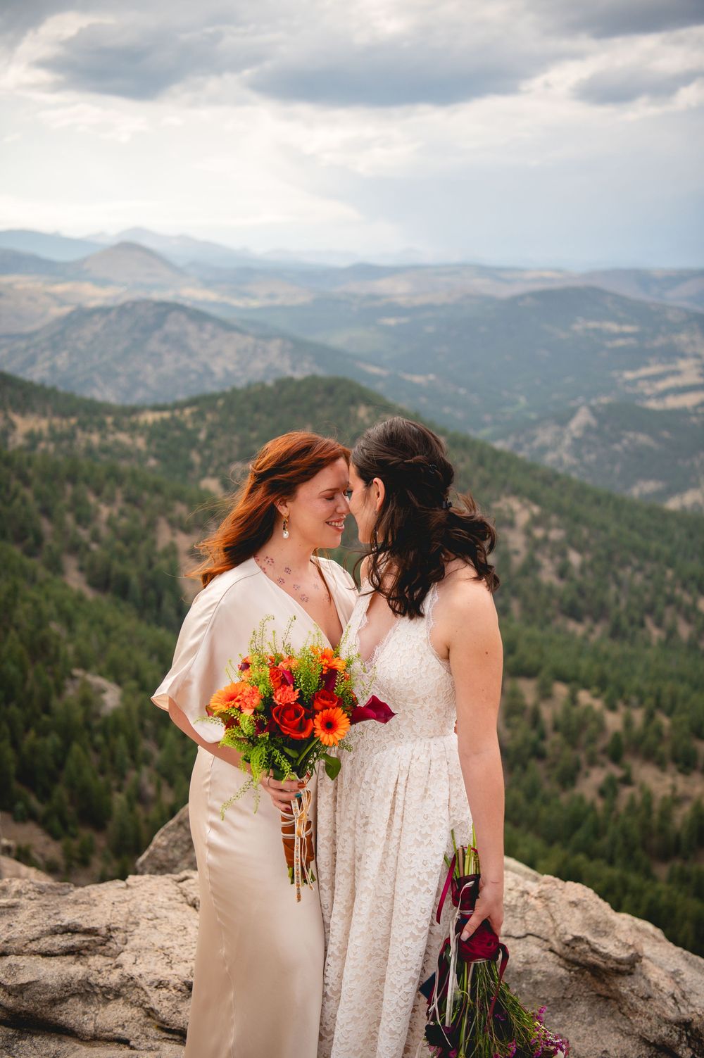 Two brides share an intimate moment on a mountain summit, one holding vibrant orange bouquet with scenic valley views behind them.