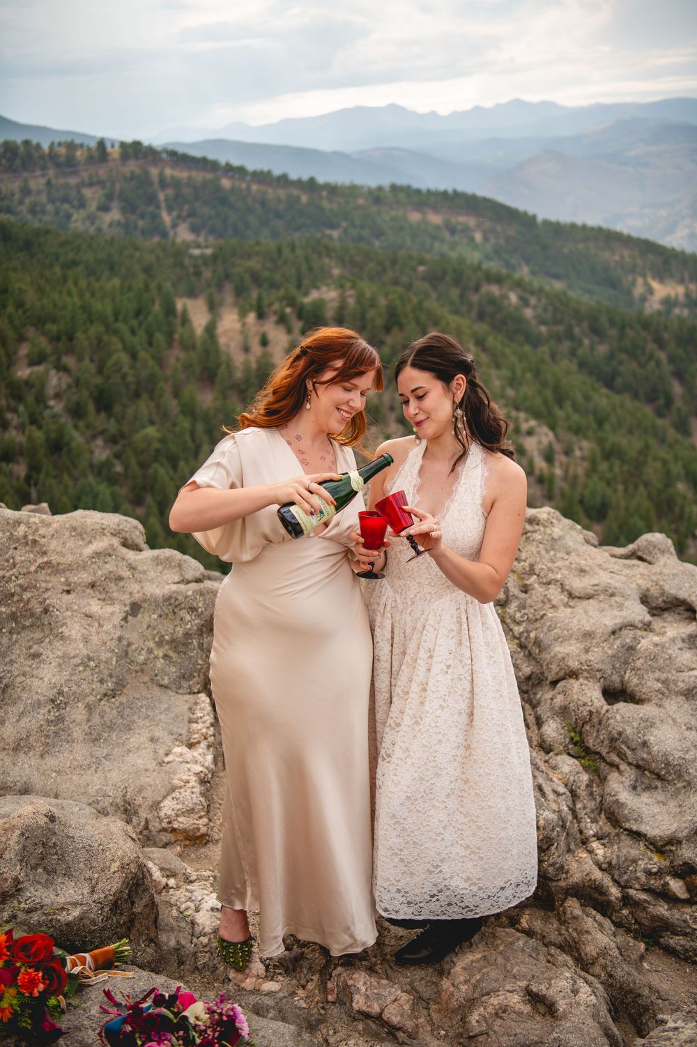 Two brides in elegant dresses toast with champagne on a rocky mountain overlook with forested peaks in the background.