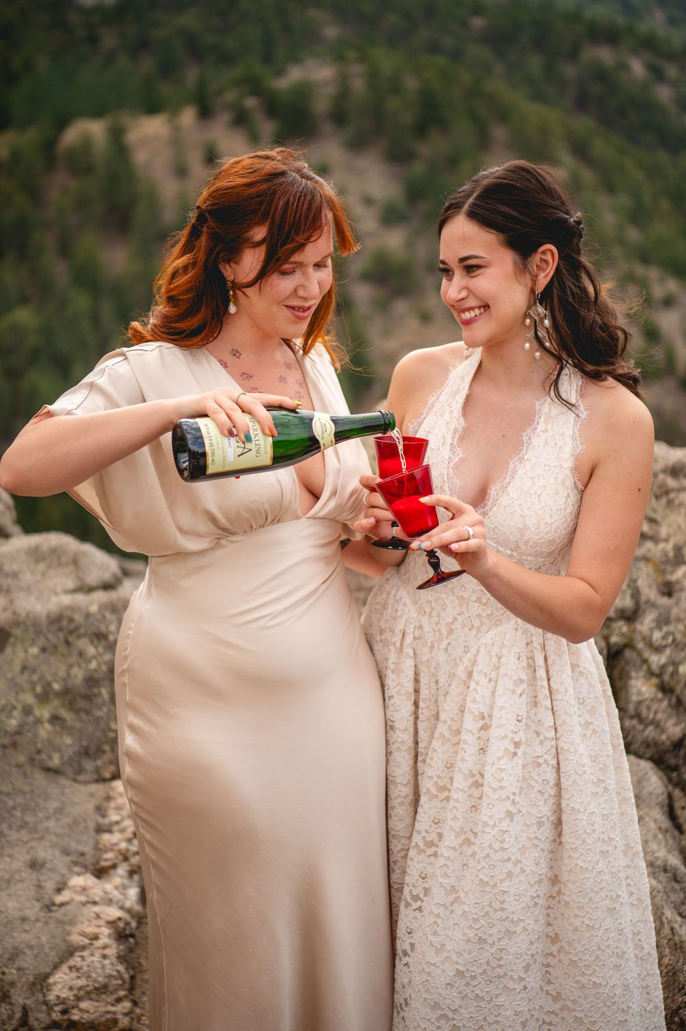 Two women in elegant dresses toast with red cups as champagne is poured outdoors with mountain views.