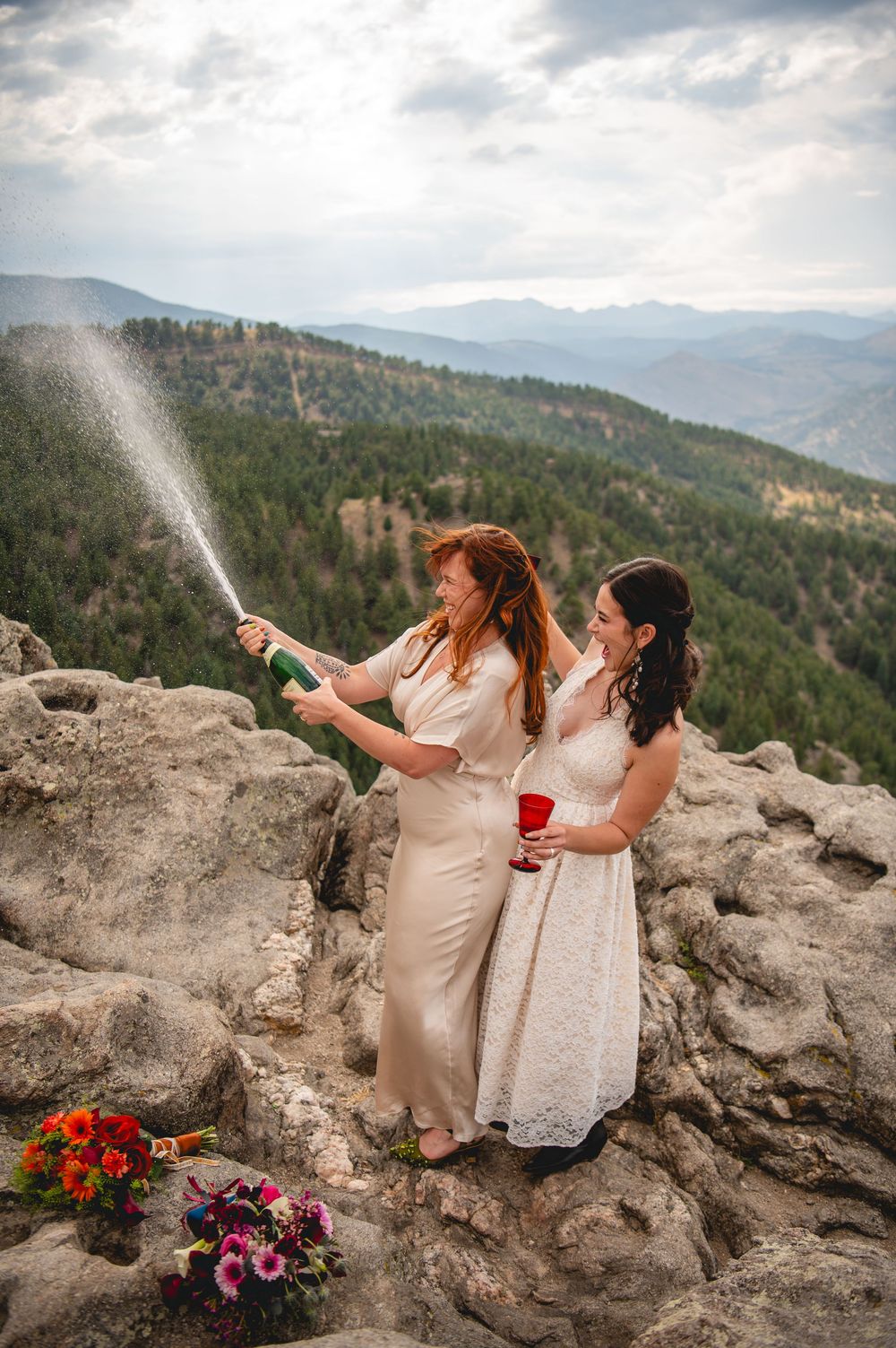 Two brides celebrate on a mountain summit by popping champagne, with scenic forested hills and cloudy skies behind them.