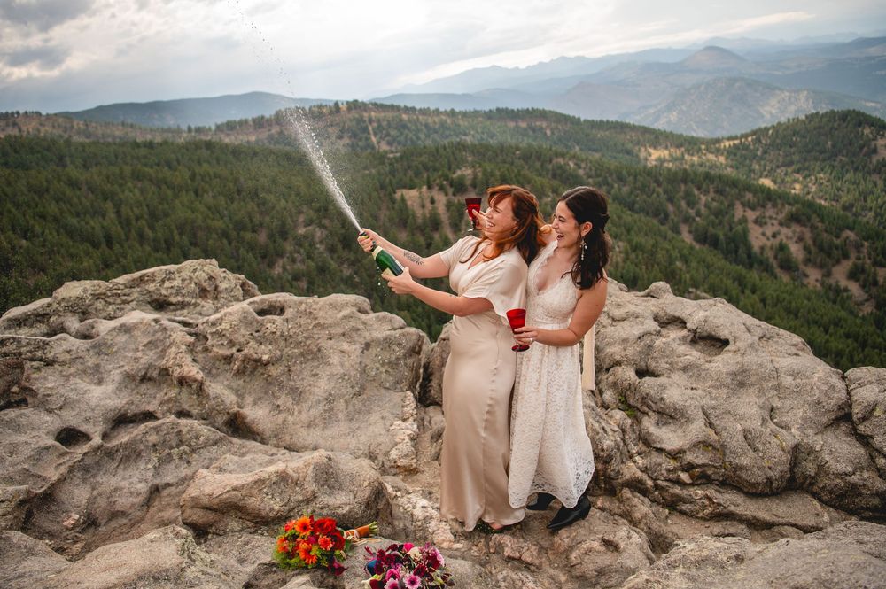 Two brides celebrate on a rocky mountain summit, popping champagne with scenic forest and mountain views behind them.