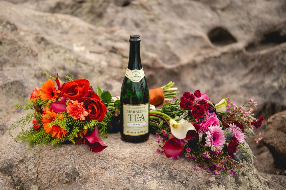 Two vibrant wedding bouquets with red and pink flowers flank a sparkling tea bottle on a rocky surface.