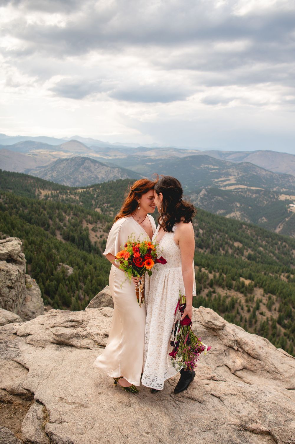 Two brides in white dresses share a romantic moment on a rocky mountain overlook with sweeping forest views.