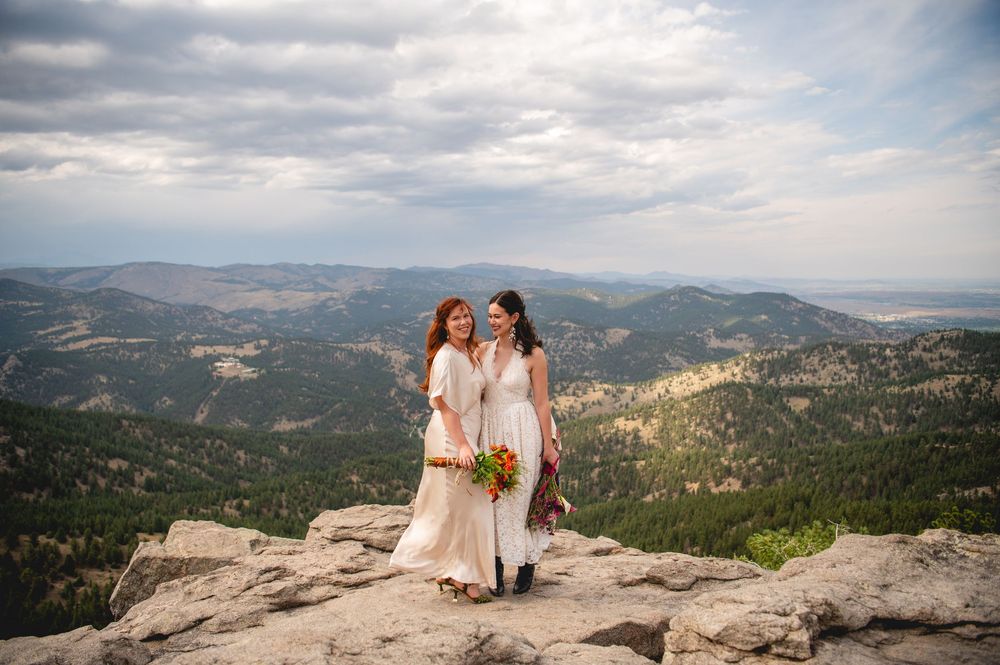 Two brides in white dresses stand on a rocky mountain summit with sweeping forest valley views on their wedding day.