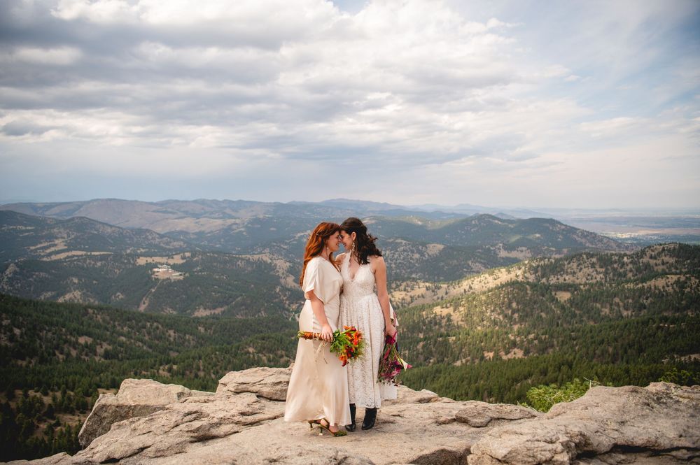 Two brides share a kiss on a rocky mountain summit with sweeping Colorado forest valley views.