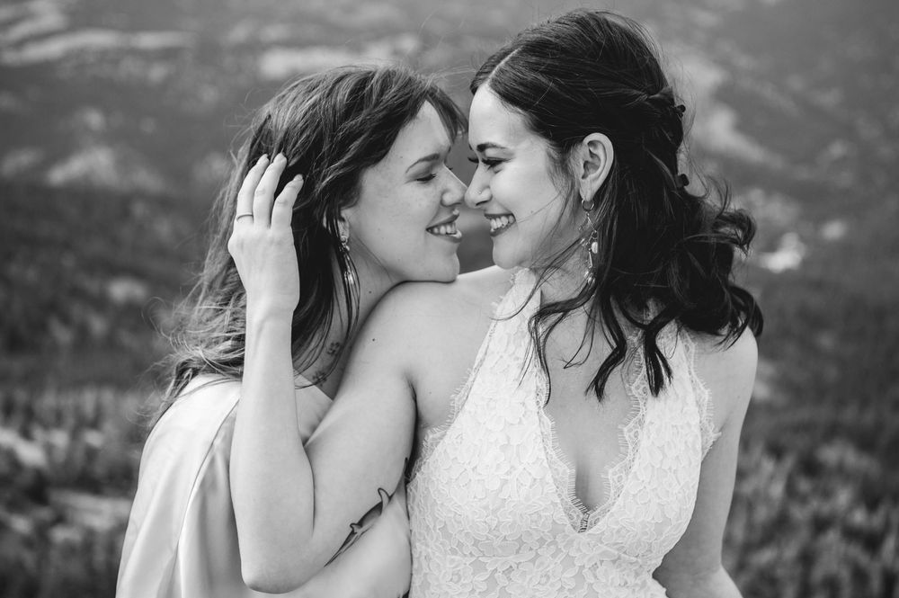 Black and white photo of two brides sharing a tender, joyful moment with mountain landscape in the background.