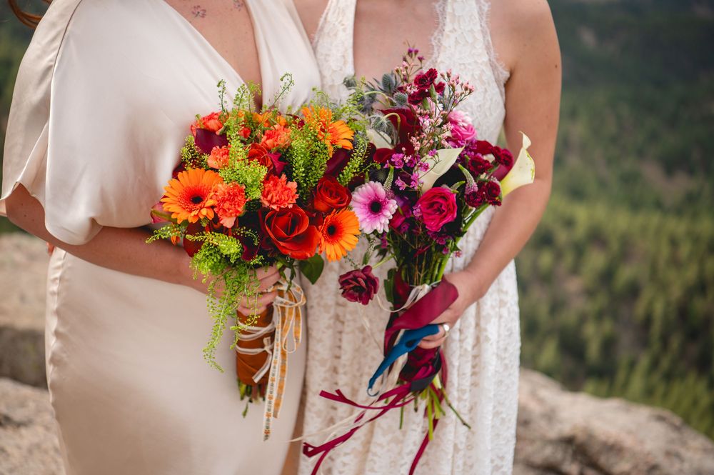 Two brides holding vibrant wedding bouquets of orange gerberas and pink flowers outdoors.