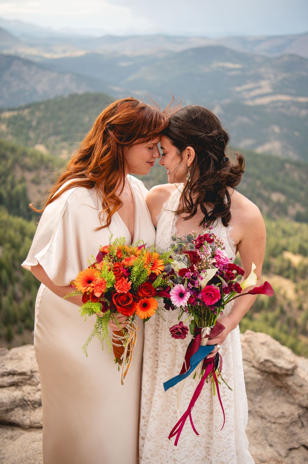 Two brides in white dresses share a romantic moment on a mountain overlook, holding vibrant colorful bouquets.