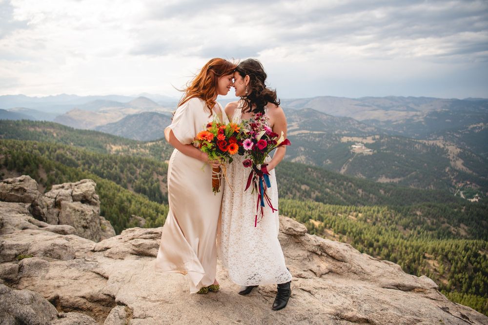 Two brides share a tender moment on a rocky mountain summit, holding colorful bouquets with a scenic valley view.