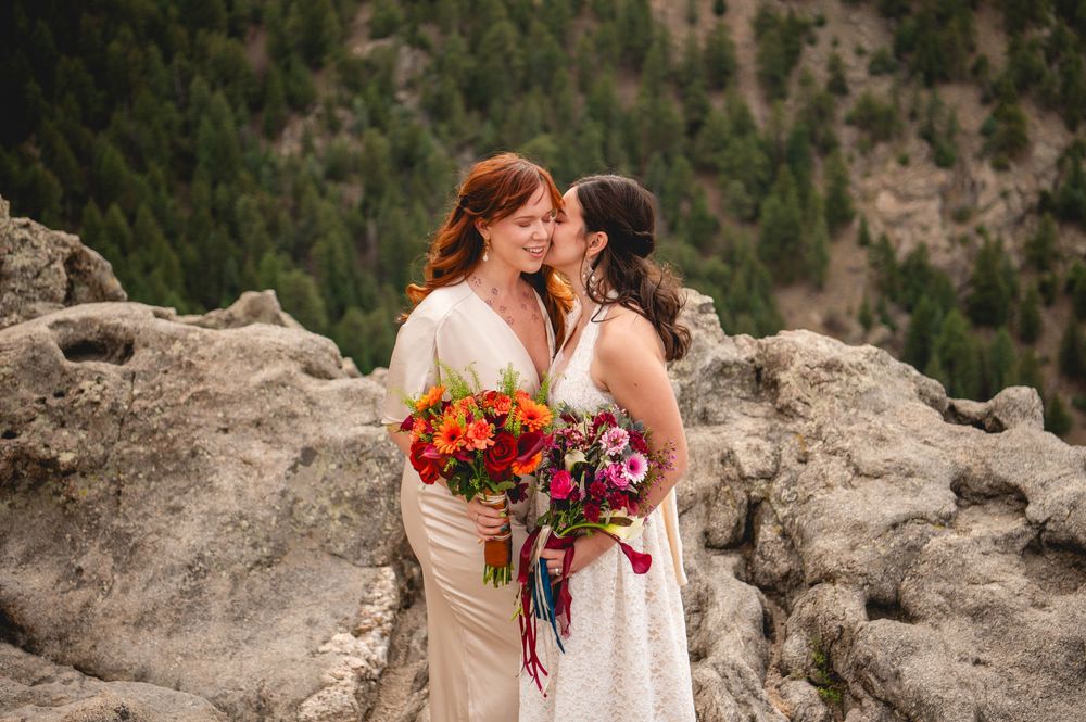 Two brides share a kiss on rocky mountain overlook, one holding vibrant bouquet of red and orange flowers.