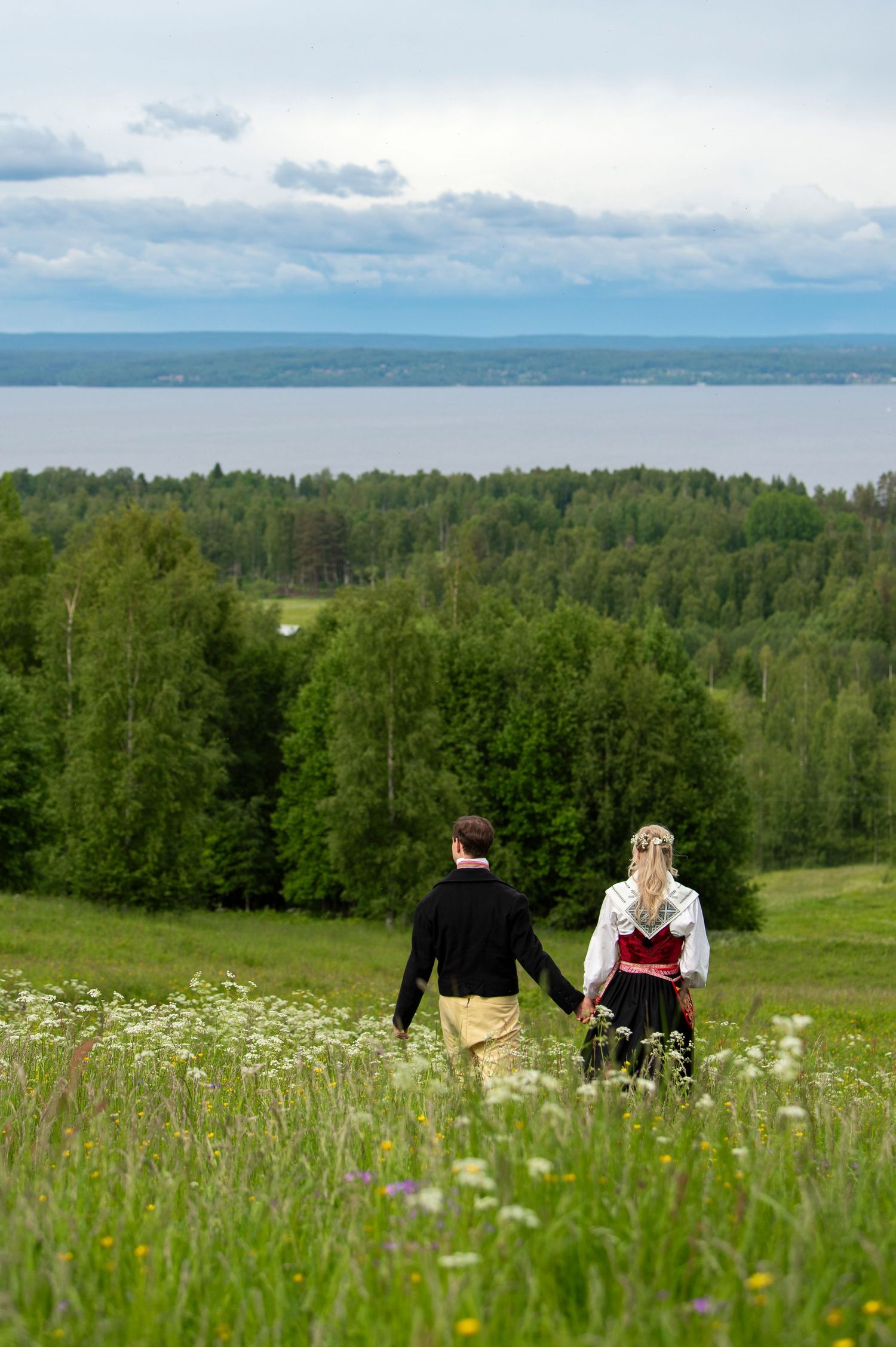 Couple in traditional attire holding hands in a wildflower meadow with forest and lake views under cloudy skies.