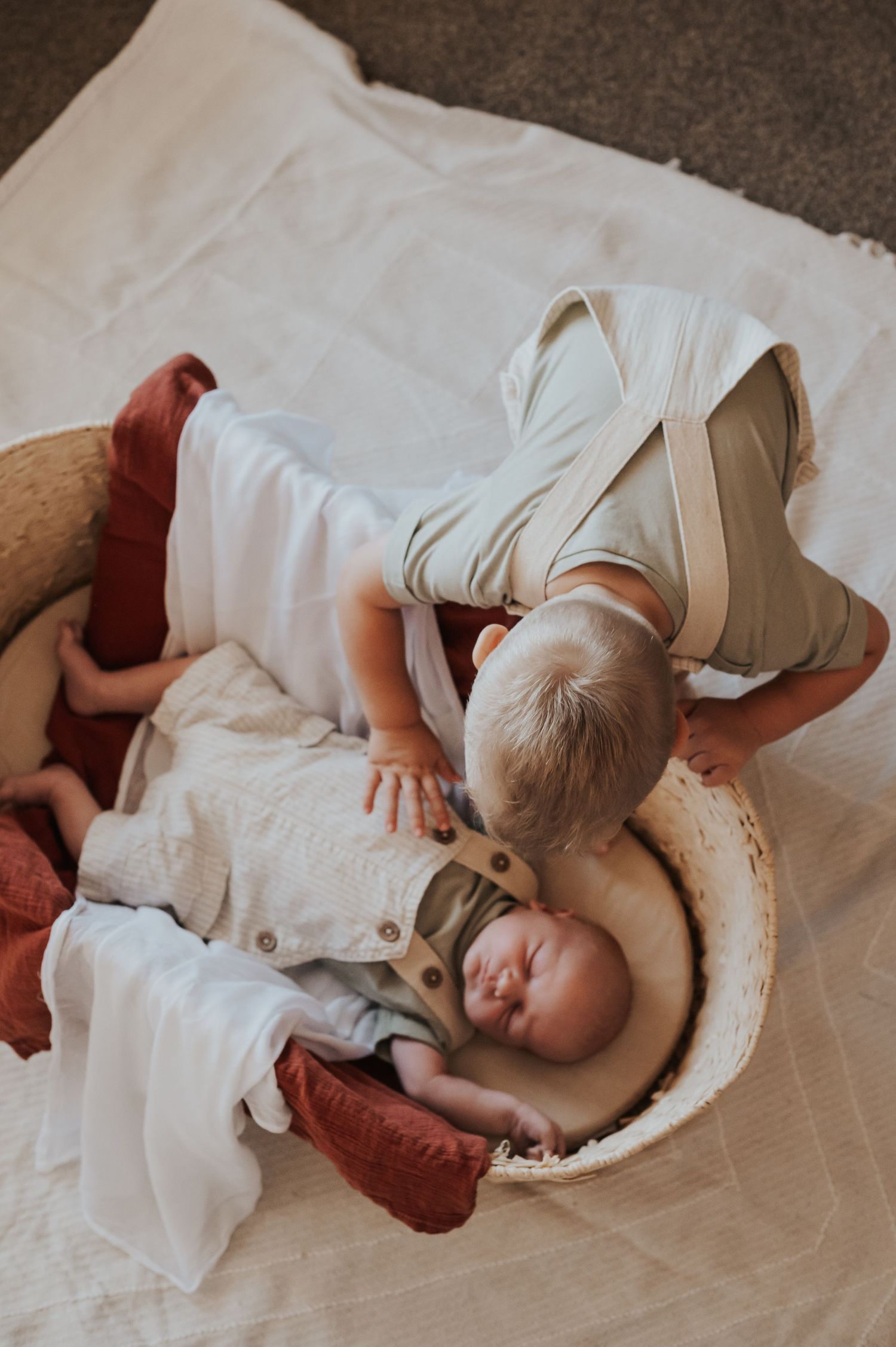 Toddler leans over wicker bassinet to look at sleeping newborn sibling on white blanket.