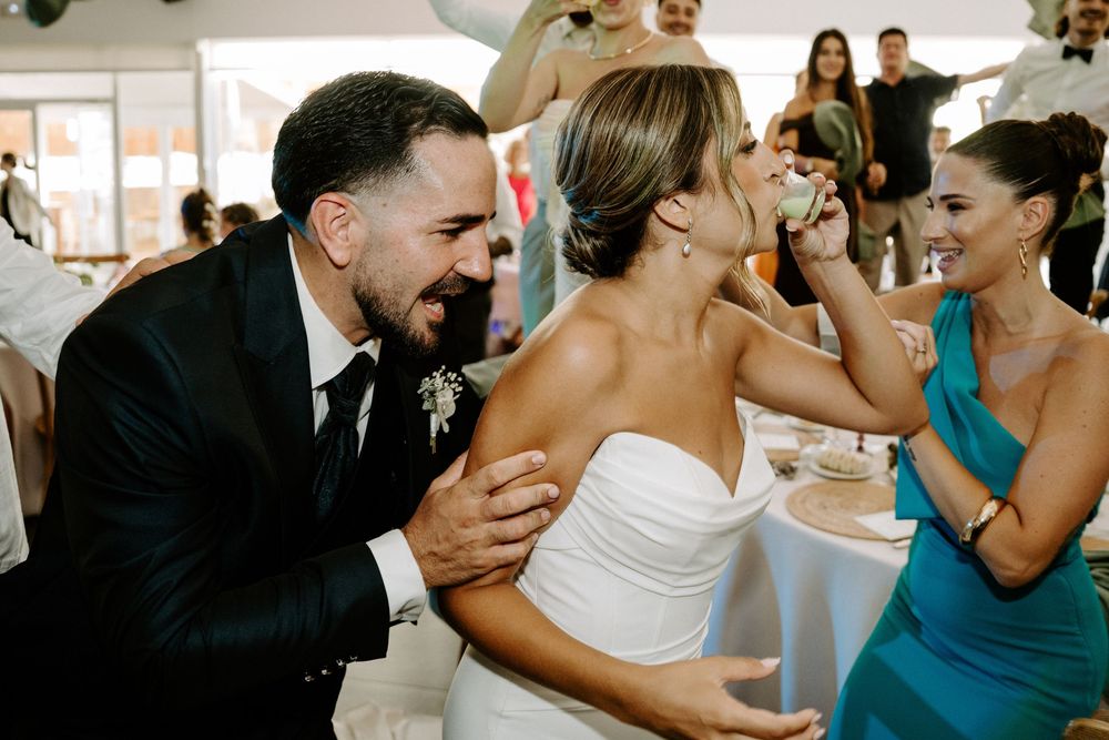 Bride and groom share a joyful moment as a bridesmaid feeds them cake at their wedding reception.