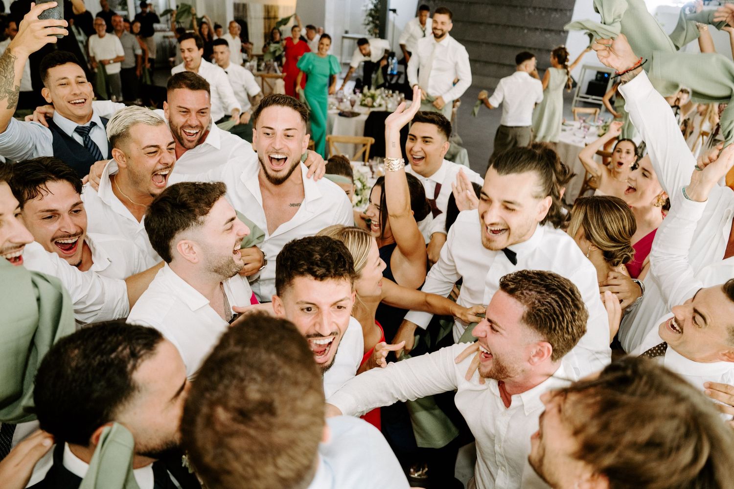 Joyful wedding reception crowd of guests in white shirts celebrating and cheering together at a lively party.