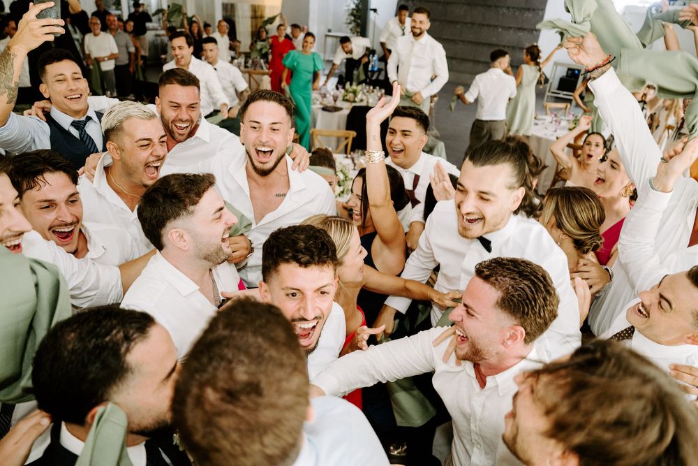 Joyful wedding reception crowd of guests in white shirts celebrating and cheering together at a lively party.