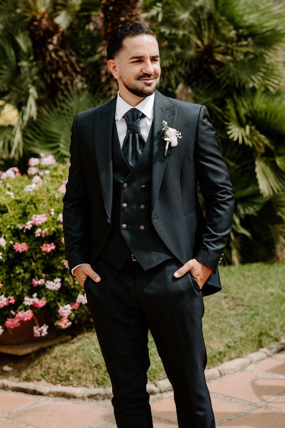 Dapper groom in black tuxedo with boutonniere poses outdoors among tropical palms and pink flowers.