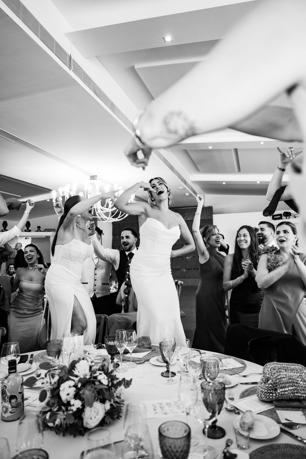 Black and white photo of two brides dancing joyfully at their wedding reception with guests cheering.