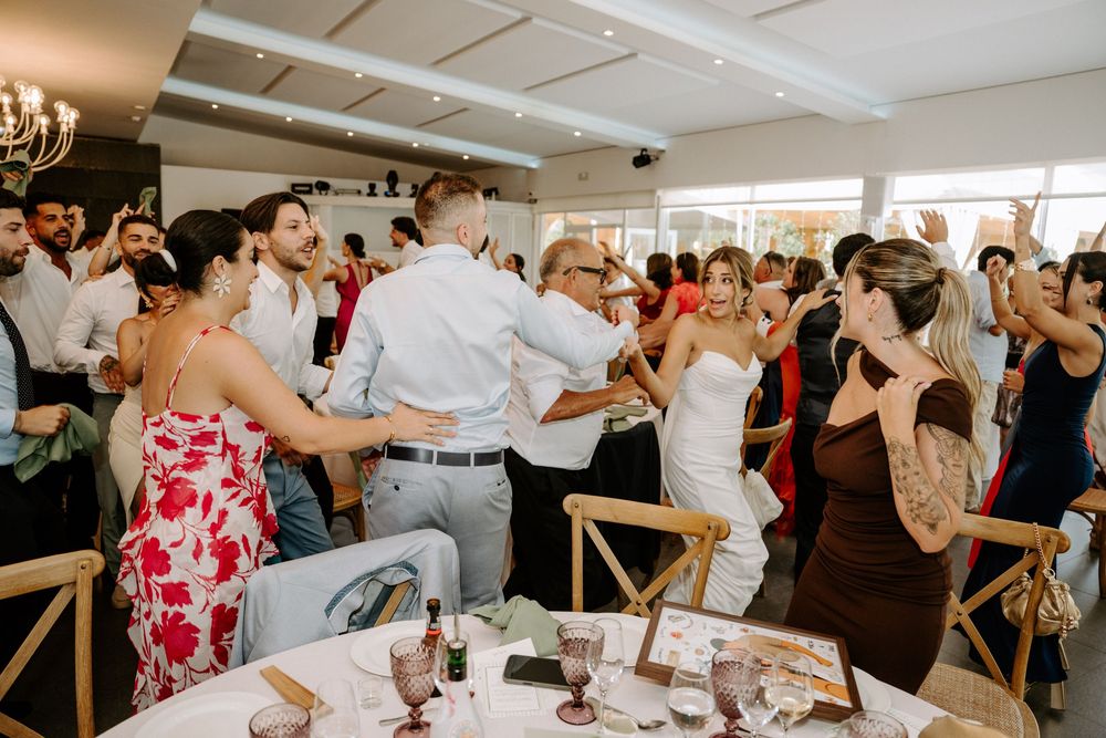 Bride and groom dancing with guests at elegant indoor wedding reception with wooden chairs.