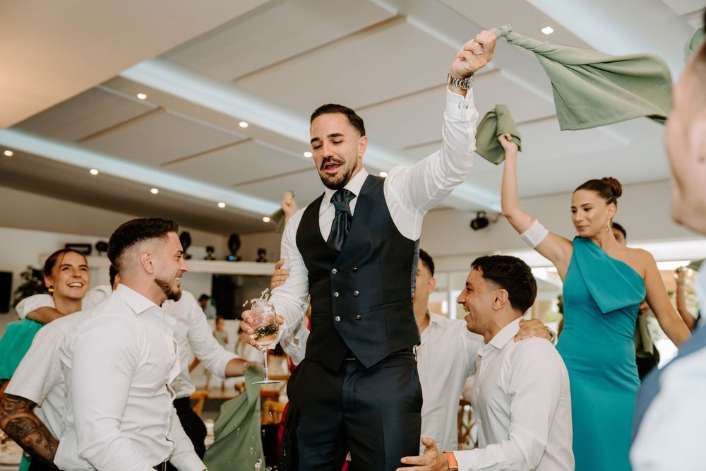 Groom dances energetically with groomsmen waving a green cloth at a lively indoor wedding reception.