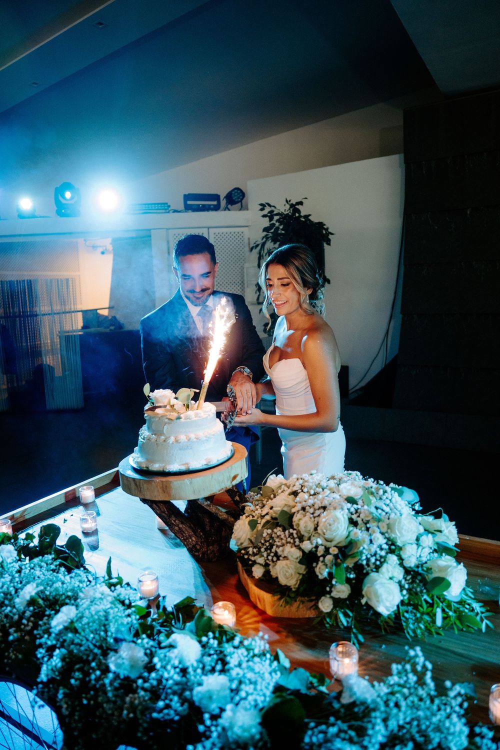 Bride and groom cutting their white wedding cake with sparkler in dramatic blue evening lighting.