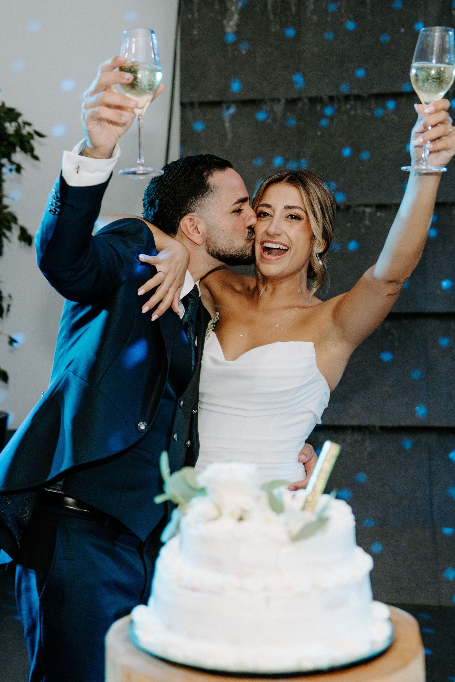 Bride and groom raising champagne glasses and kissing beside white wedding cake with blue confetti falling.