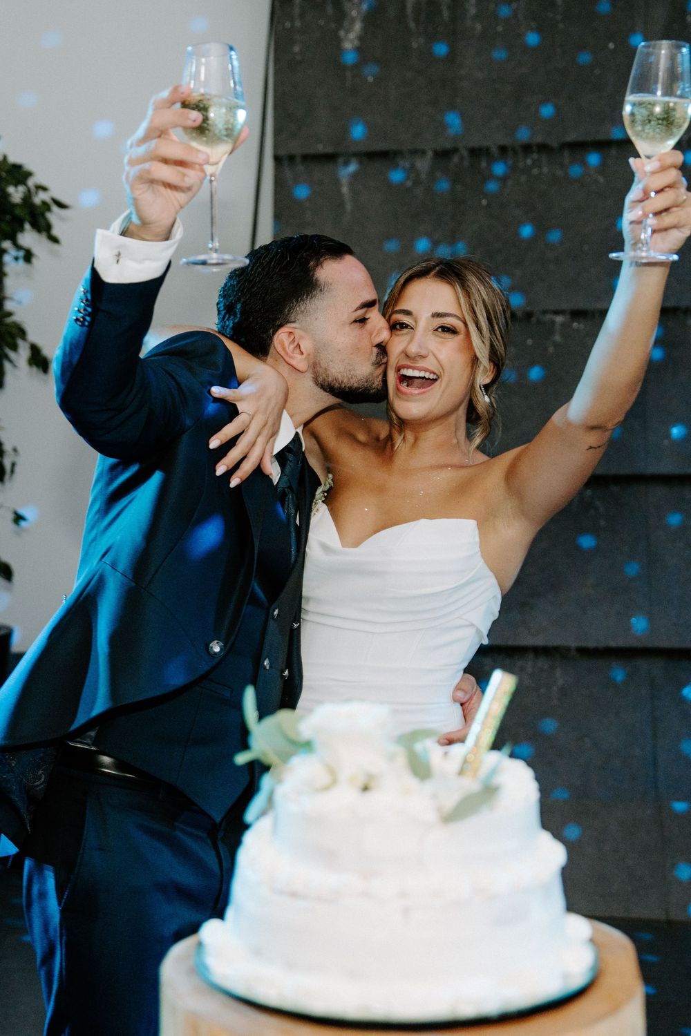 Bride and groom raising champagne glasses and kissing beside white wedding cake with blue confetti falling.