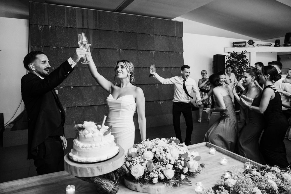 Black and white photo of bride and groom toasting with champagne glasses next to their wedding cake.
