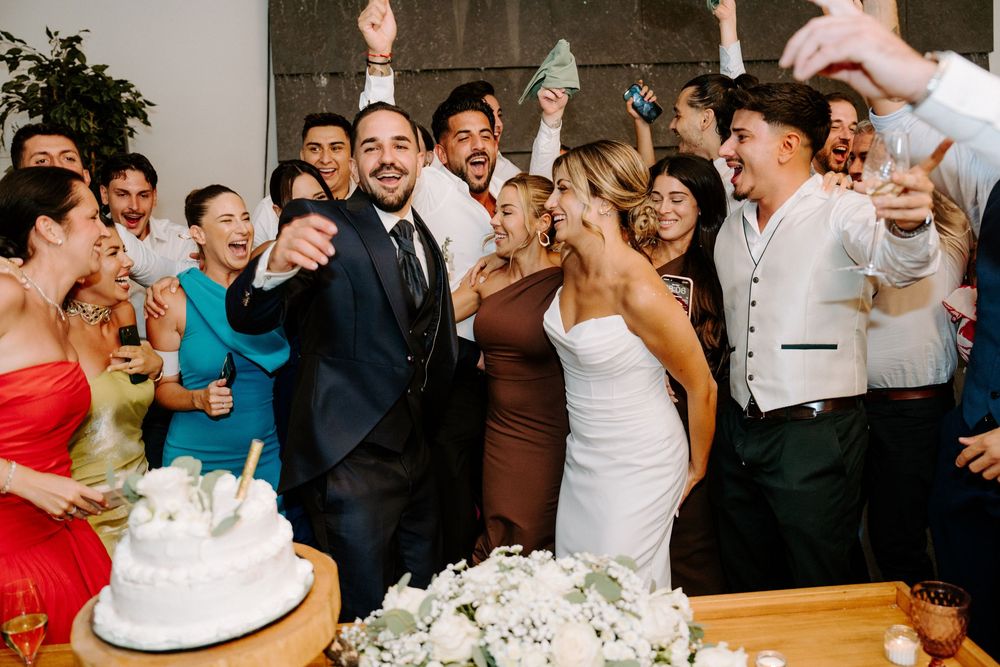 Bride and groom cutting wedding cake surrounded by cheering guests at reception.