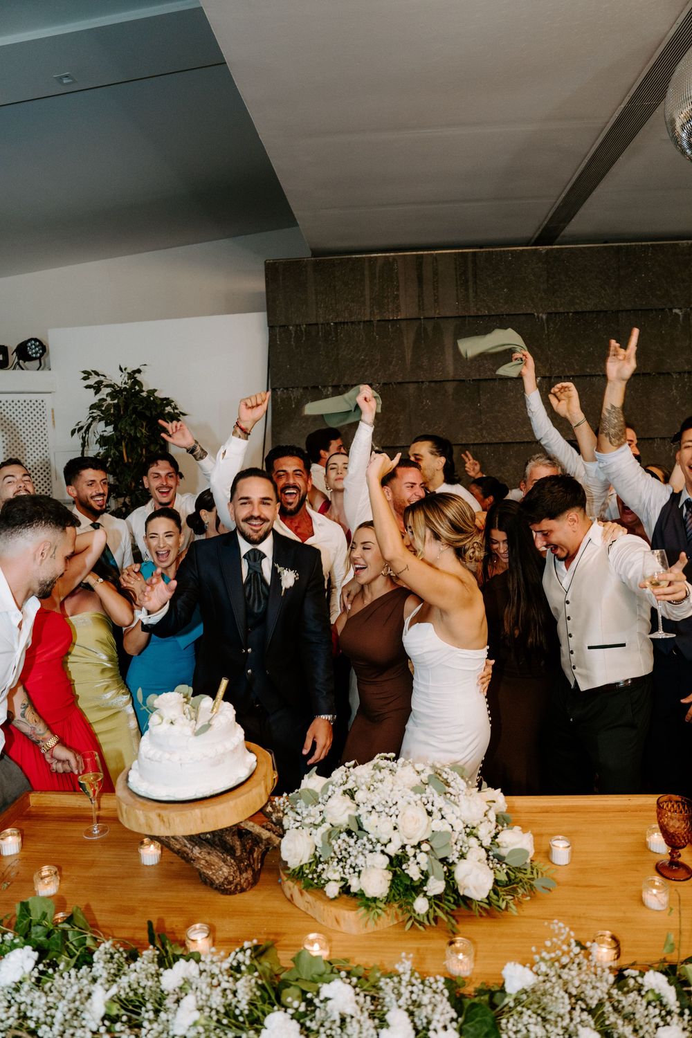 Bride and groom celebrate cake cutting surrounded by cheering guests at indoor wedding reception.