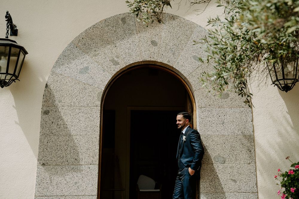 Groom in navy suit standing elegantly by a stone arched doorway with lush greenery.
