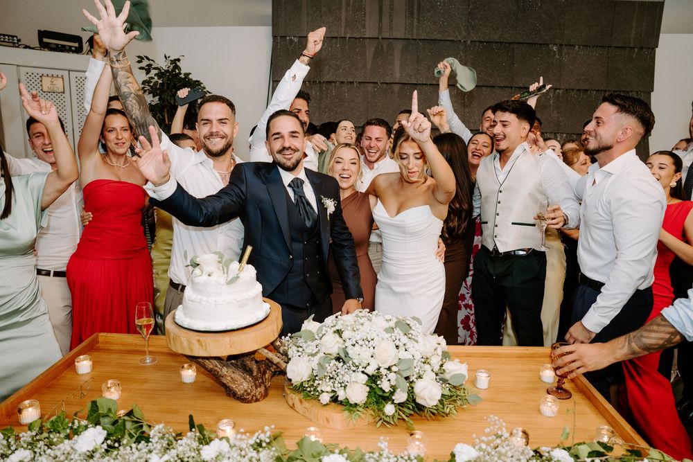 Smiling newlyweds cut white wedding cake as joyful guests celebrate around decorated reception table.