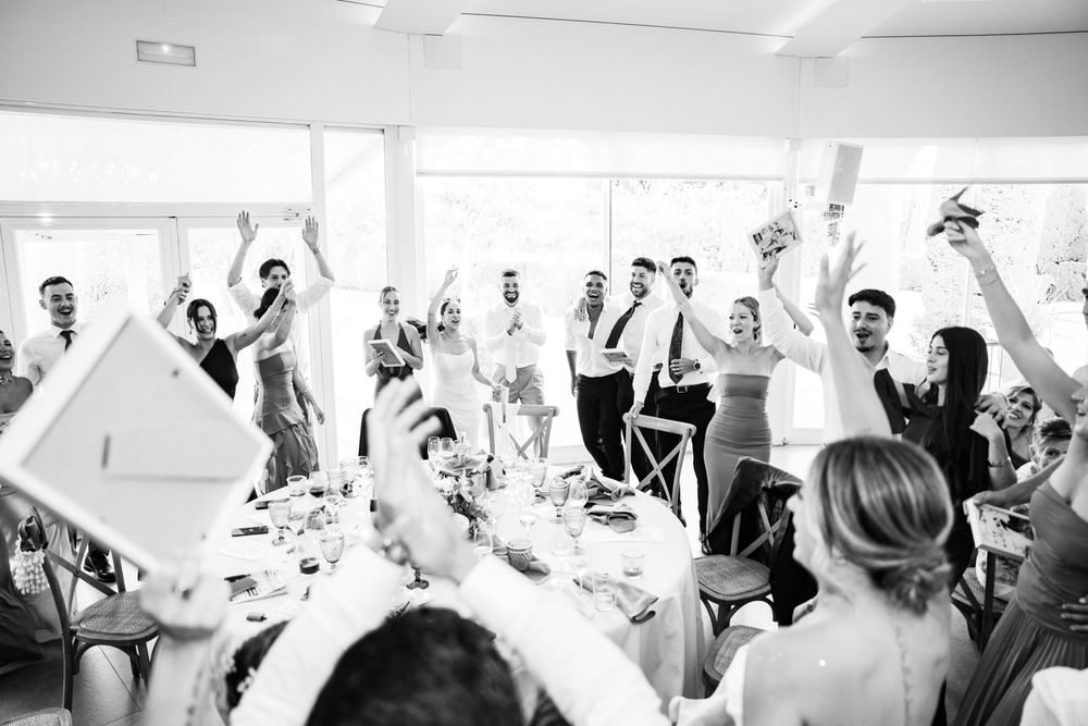 Black and white photo of wedding guests cheering and raising paddles during lively reception celebration.