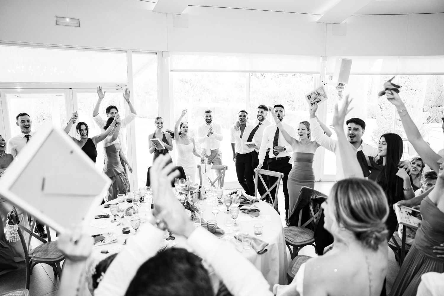 Black and white photo of wedding guests cheering and raising paddles during lively reception celebration.