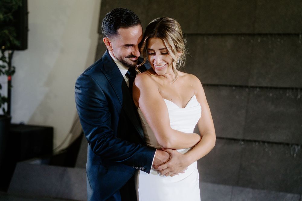 Groom in navy suit tenderly embraces smiling bride in white gown during intimate wedding moment.