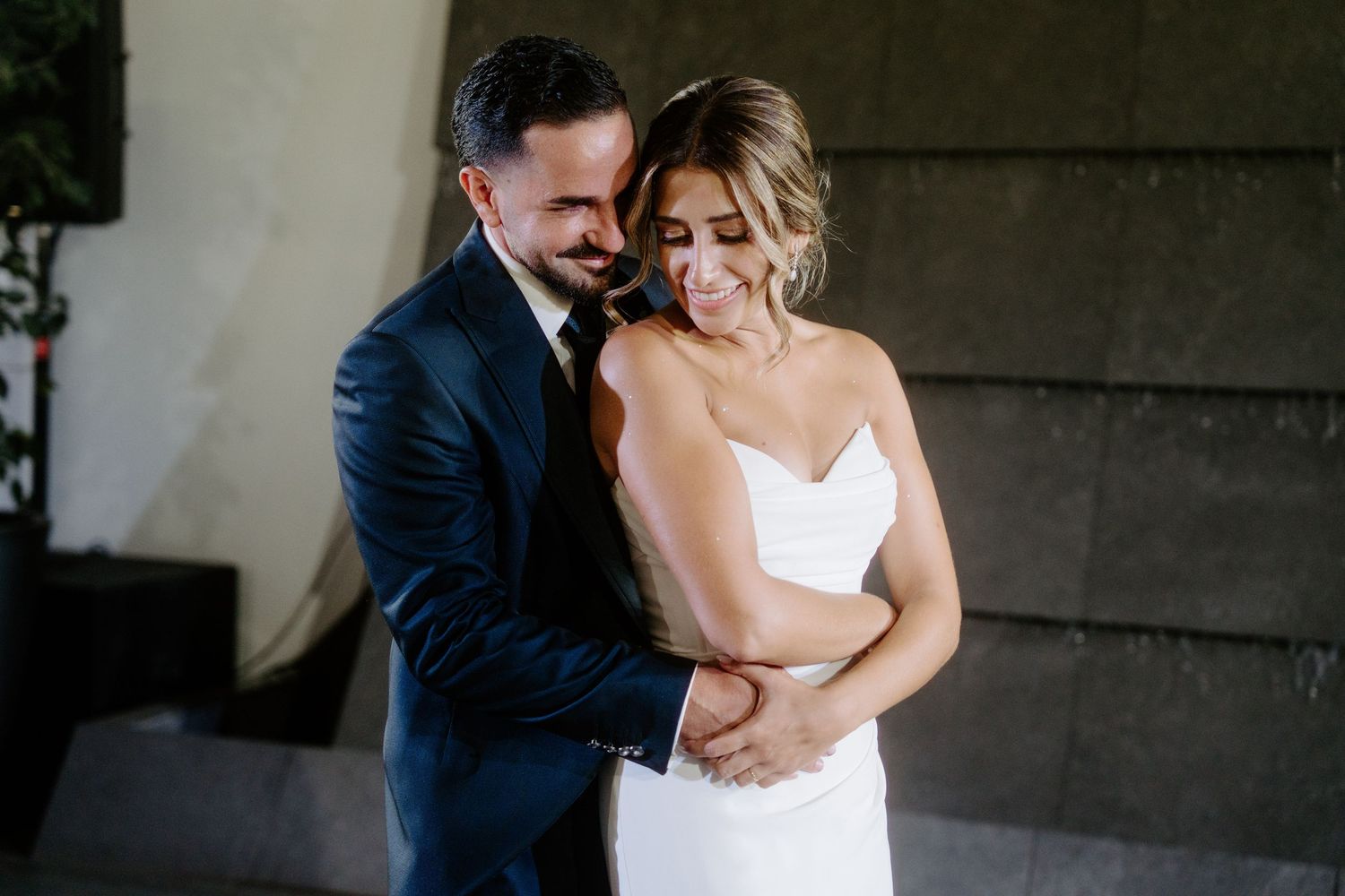 Groom in navy suit tenderly embraces smiling bride in white gown during intimate wedding moment.