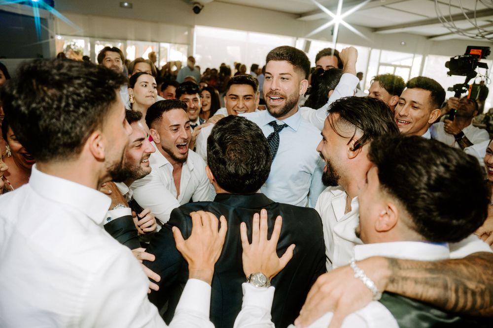 Groom lifted and celebrated by group of cheering male friends on wedding reception dance floor.