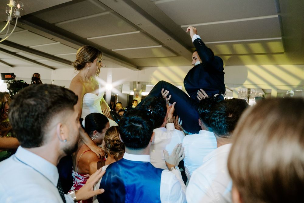 Groomsmen lifting groom into the air as bride watches during energetic indoor wedding reception.