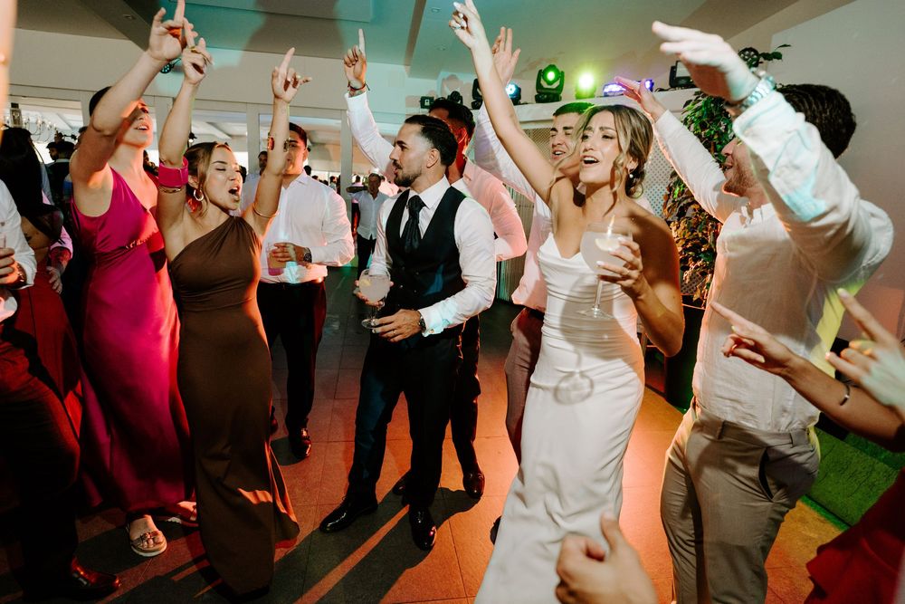Bride and groom dancing joyfully with wedding guests at a lively reception party with colorful lights.