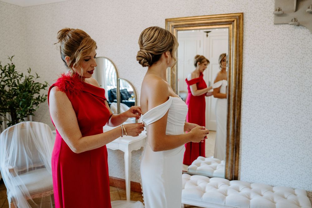 Bridesmaid in red dress helping bride in white gown prepare in front of gold mirror on wedding day.
