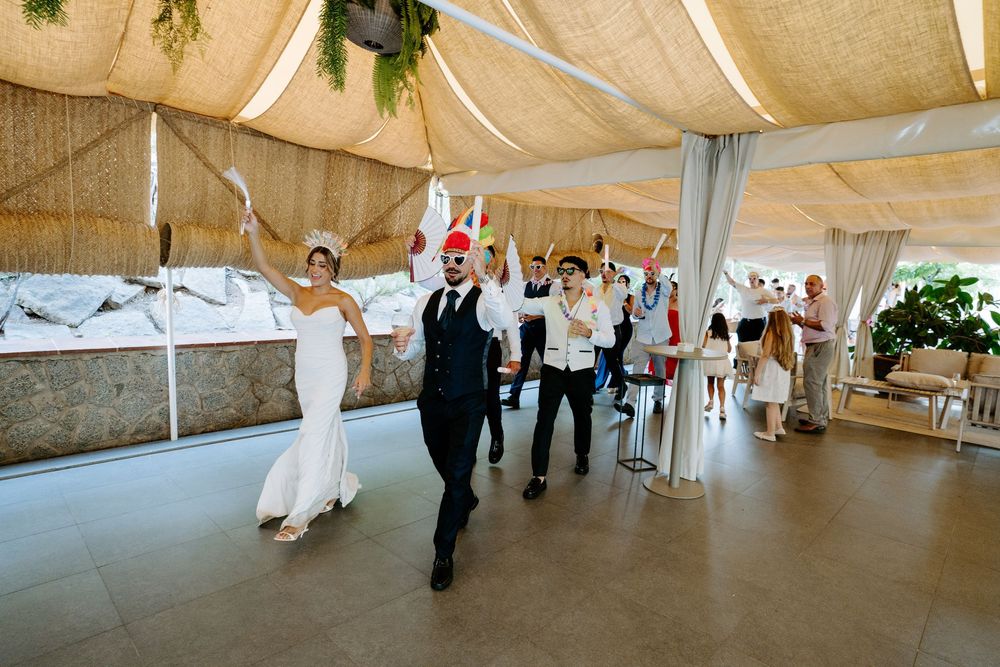 Bride and groom lead a festive wedding party entrance under a draped tent with guests celebrating.