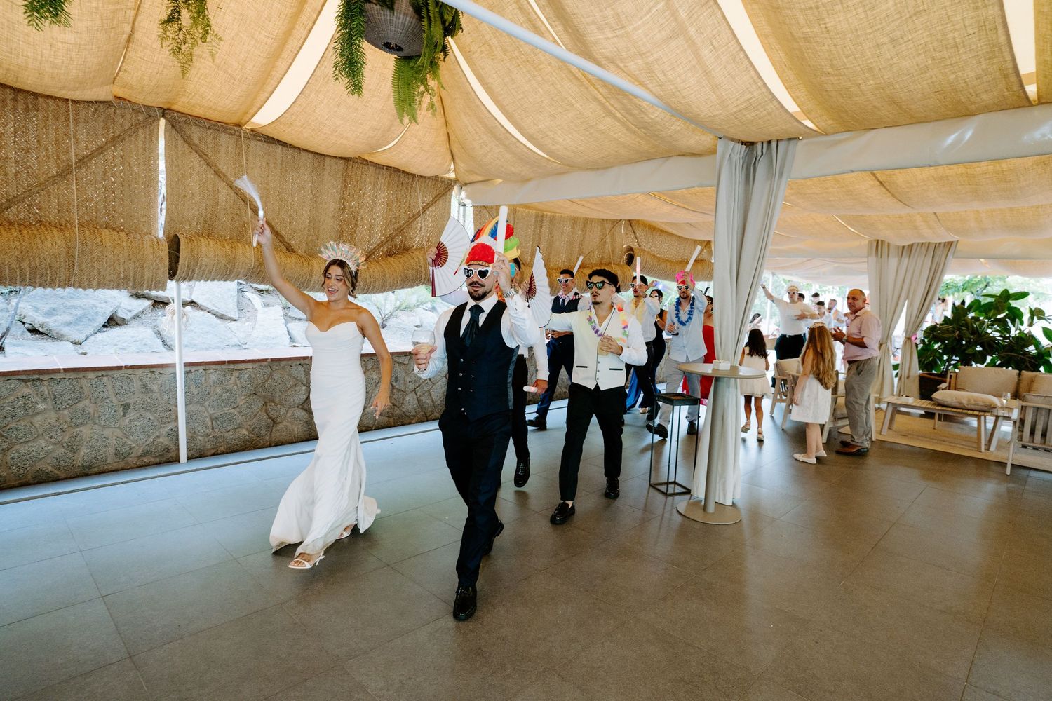 Bride and groom lead a festive wedding party entrance under a draped tent with guests celebrating.